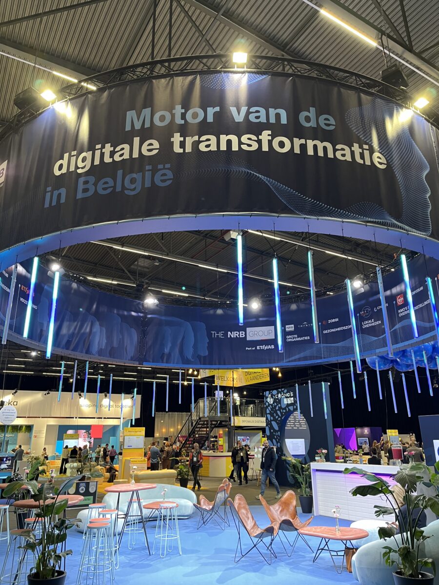 Interieur van een technologie- of digitaliseringsbeurs in België, met bezoekers, informatiestands en een centrale ontmoetingsruimte.